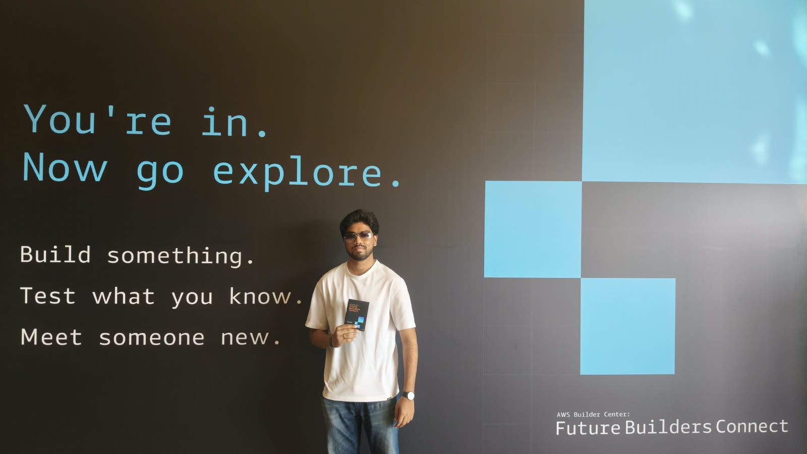 Victor at the AWS Builder Center wall that reads: You're in. Now go explore.