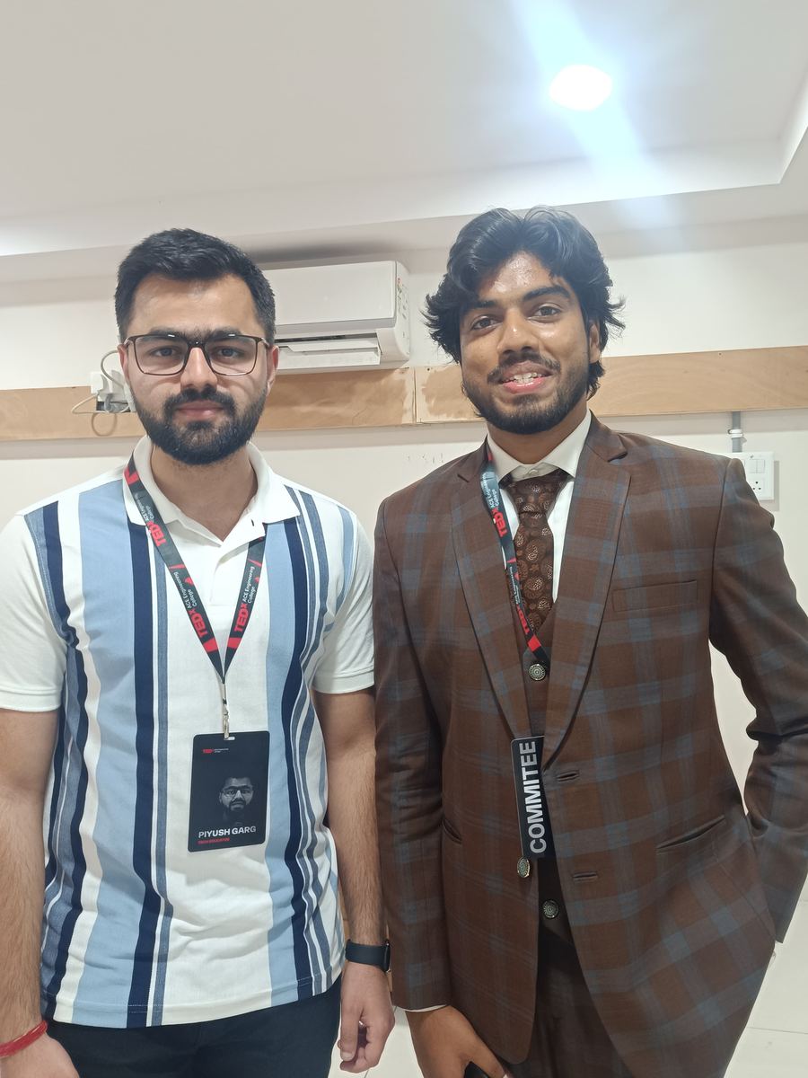 Victor in a checked brown suit with a committee lanyard beside Piyush Garg